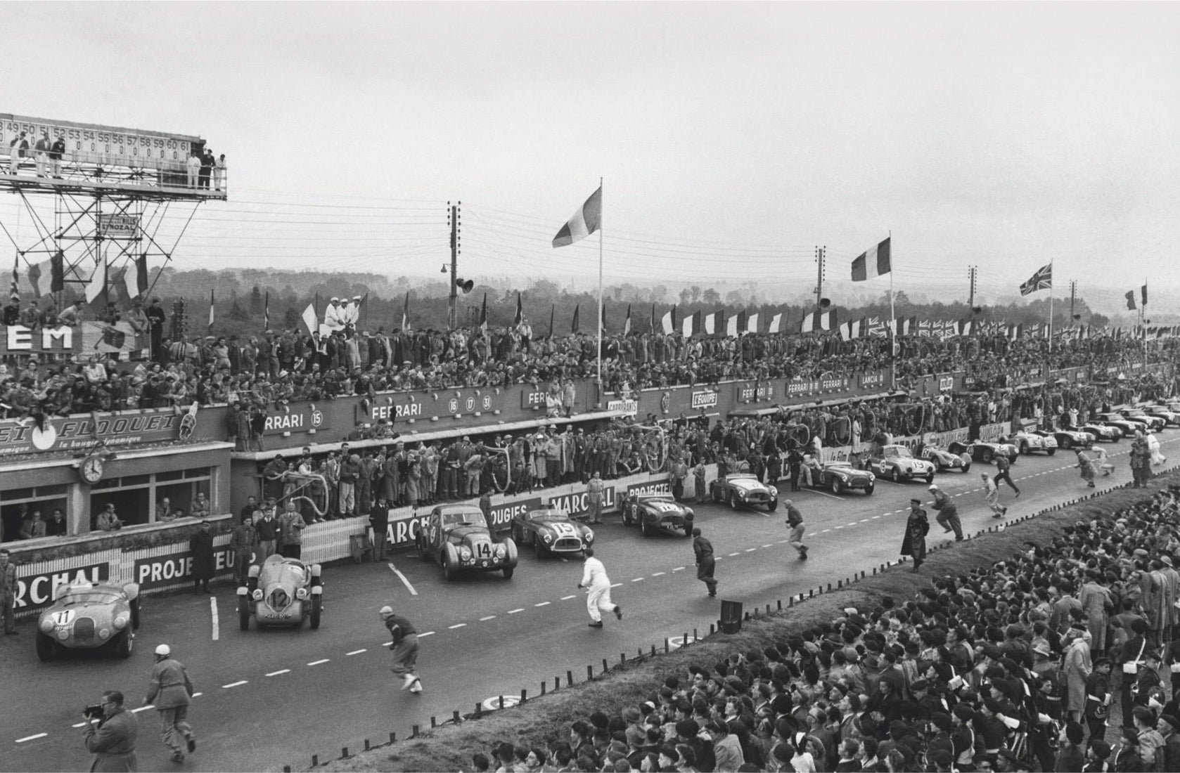 Vintage black and white photo of classic 24 Hours of Le Mans race start, with cars lined up on the track and drivers running towards their vehicles, cheered by a large crowd.