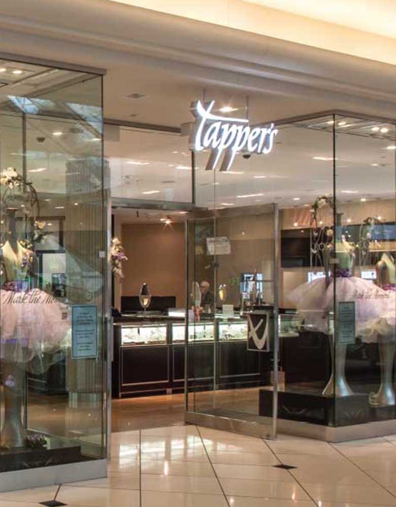 Glass-fronted Tapper’s jewelry boutique inside a shopping mall, with illuminated display cases and mannequins dressed in pastel tulle gowns visible through the glass.