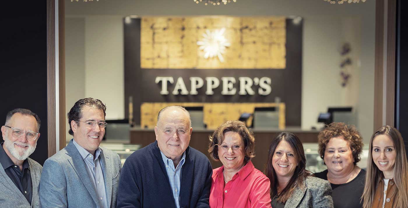 Group photo of seven smiling staff members standing together in front of Tapper's jewelry store with elegant interior and display cases