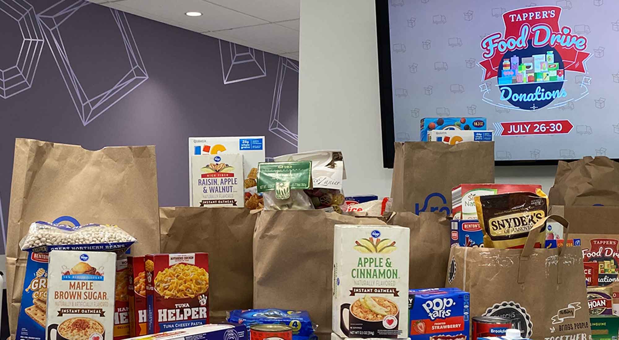Food donation collection display with brown paper bags filled with non-perishable items including oatmeal, pasta, and snacks for Tapper's Food Drive charity event July 26-30