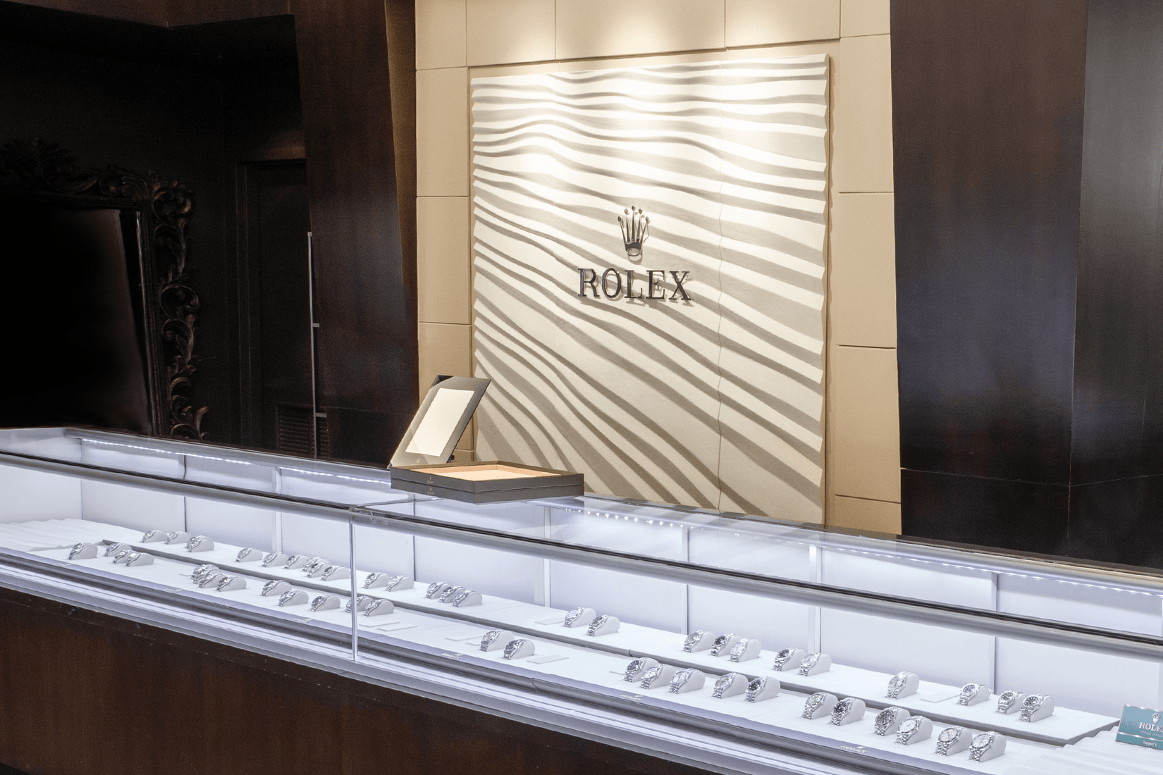 Glass-topped display case filled with rows of Rolex watches on white stands, set against a textured wall panel featuring the Rolex crown logo and name.