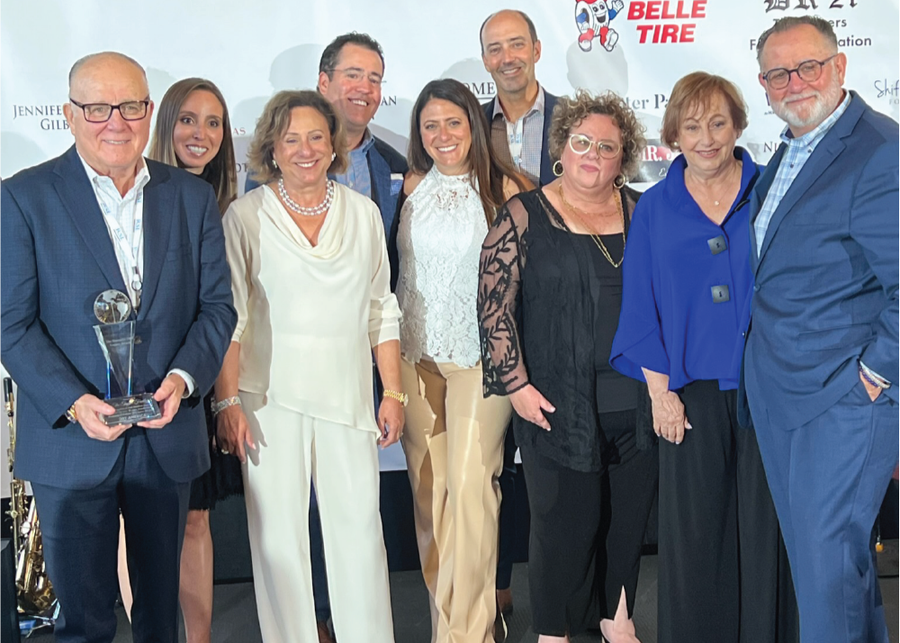 Group of ten professionally dressed people at awards ceremony, with man in blue suit holding crystal trophy, standing in front of sponsor backdrop