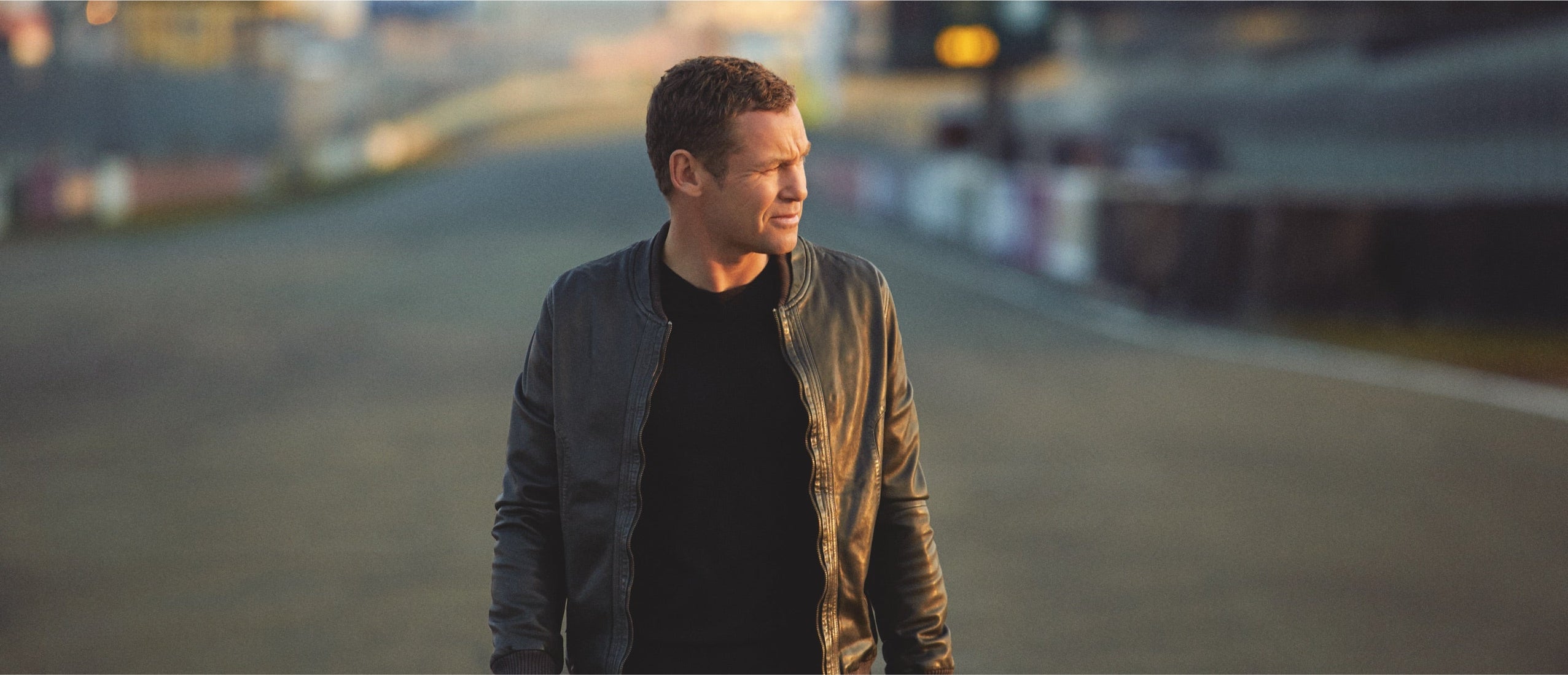Man in a leather jacket walking along an empty racetrack at dusk.