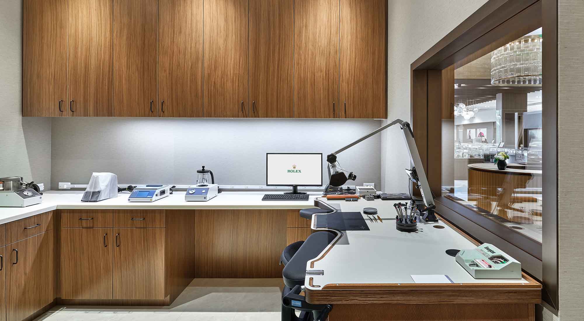 Modern watch repair workshop with walnut wood cabinetry, white countertops, precision instruments, microscope, and computer workstation for horological services