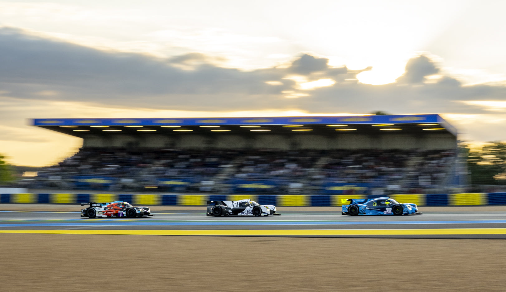 Three colorful racecars speeding around a blue and yellow track at Le Mans