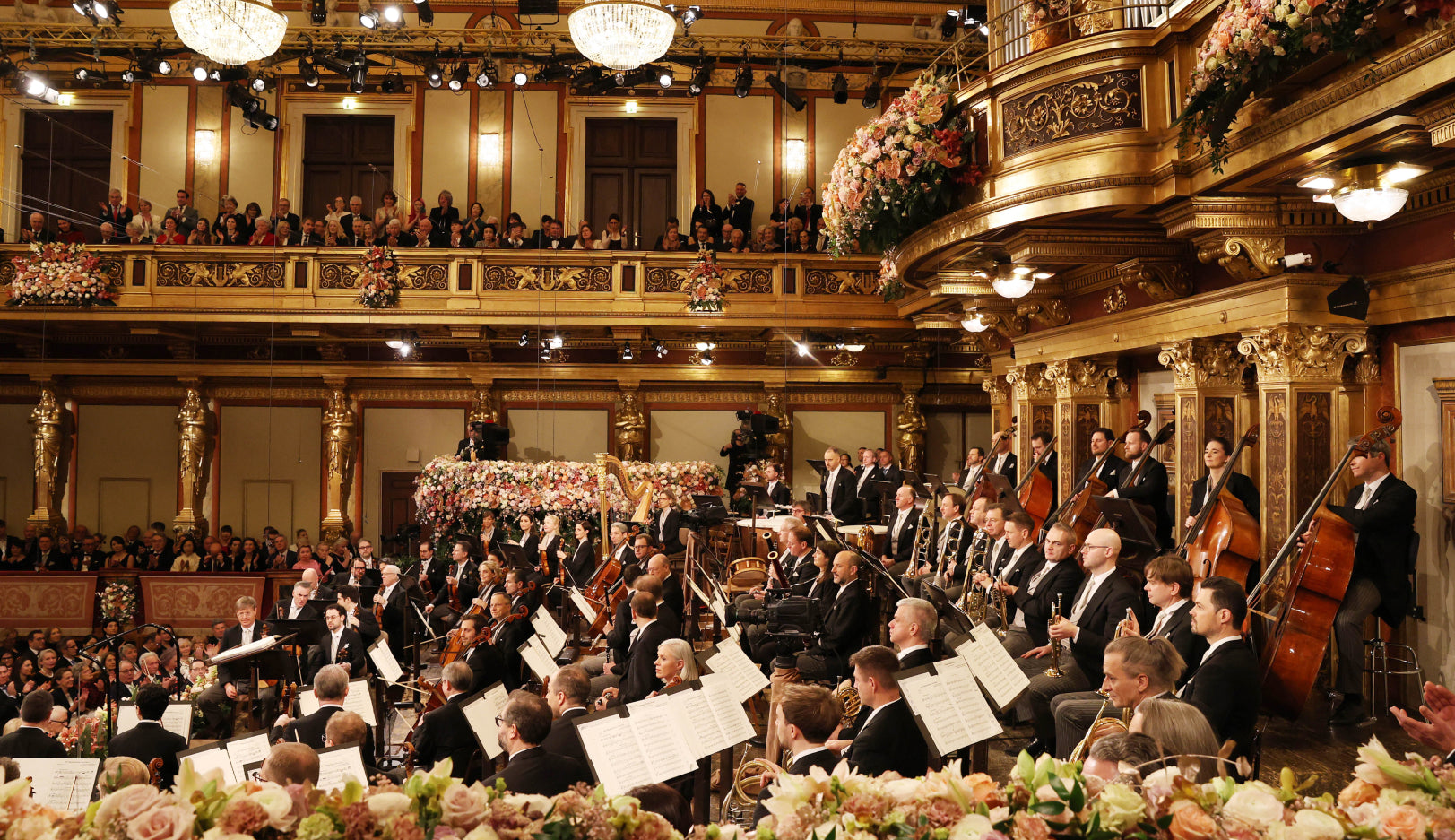 Side view of Vienna Philharmonic musicians ready to play in ornate golden hall with white and pink rose decor and chandelier lighting