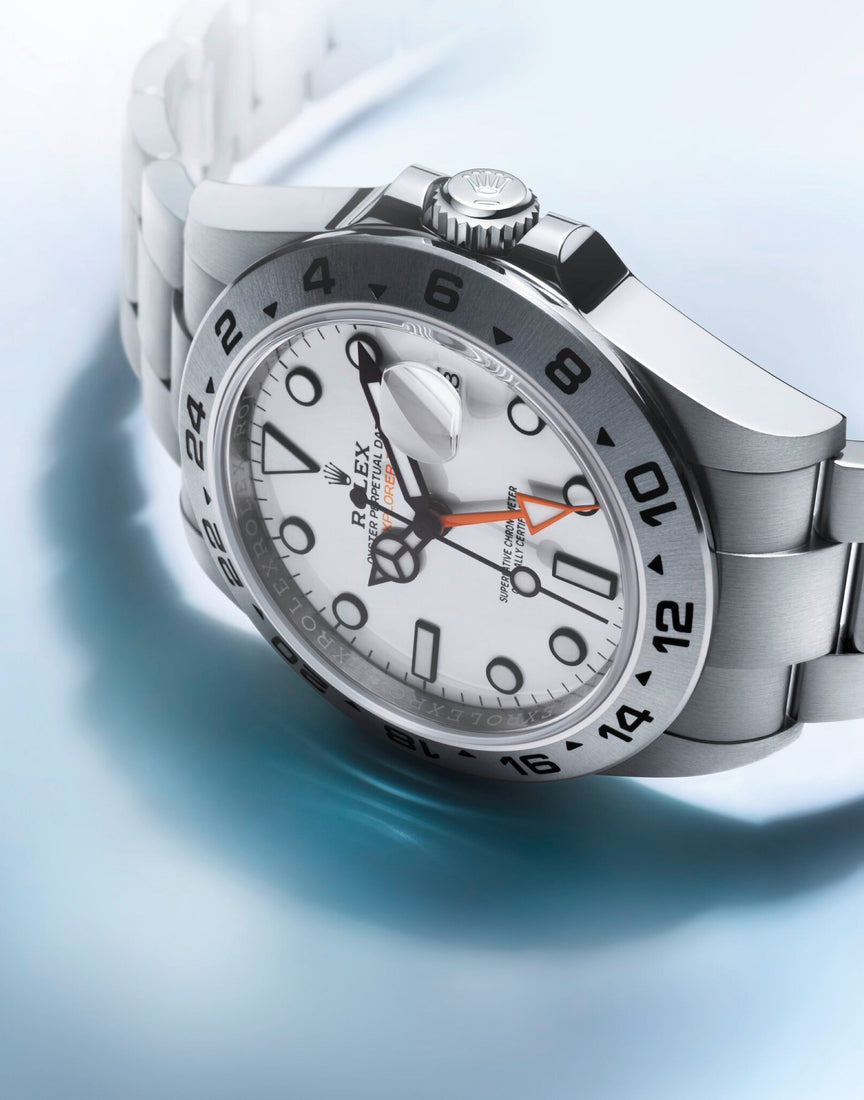 Close-up of a Rolex Explorer II watch with a white dial, black hour markers, 24-hour bezel, and stainless steel bracelet, shown resting on a light blue surface.