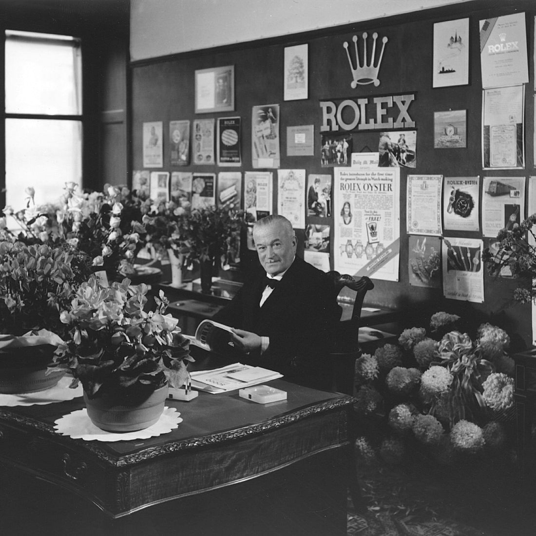 Historic black and white photo of a man seated at a desk adorned with flowers, in an office decorated with vintage Rolex advertisements and posters covering the wall.