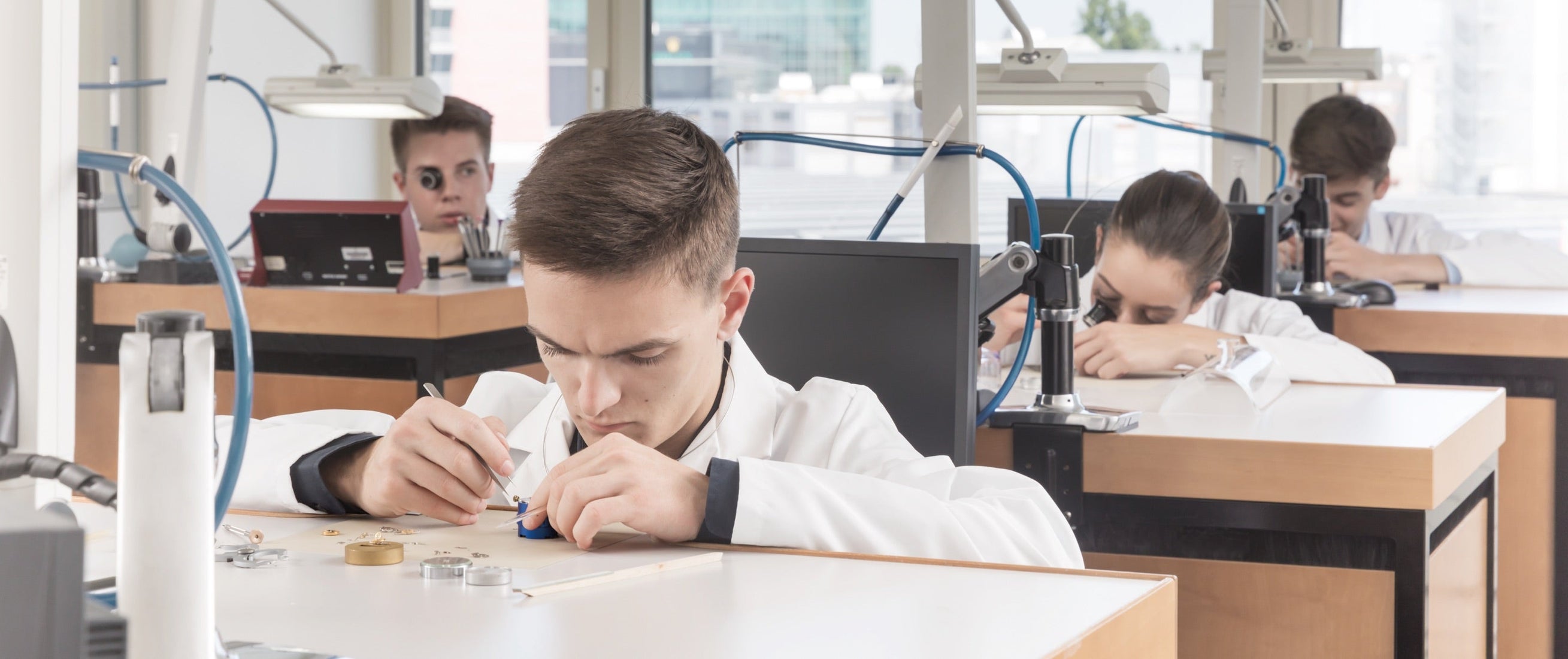 Group of technicians in white lab coats seated at workstations, assembling and inspecting watch components in a brightly lit horology workshop.