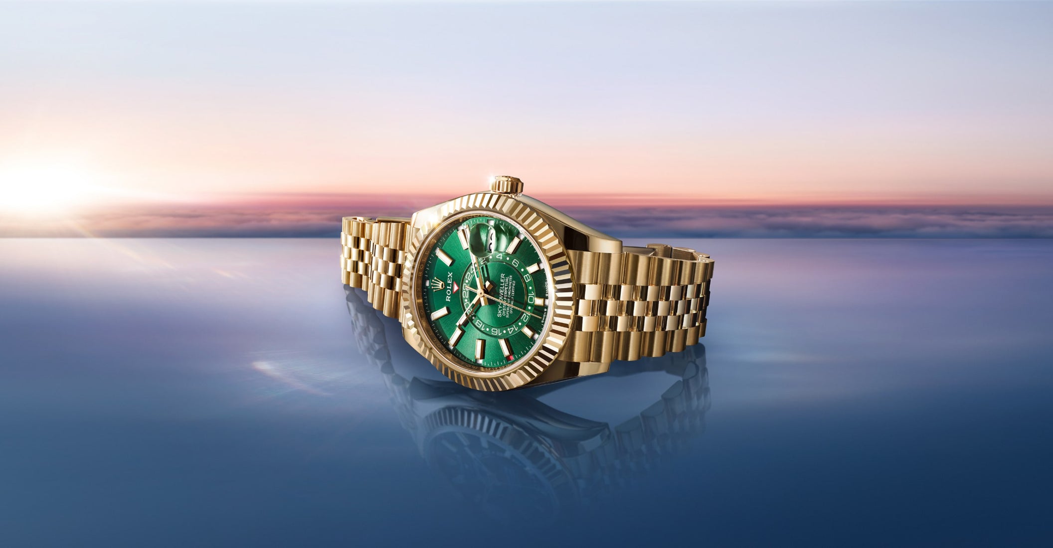 Gold Rolex watch with green dial and fluted bezel on Jubilee bracelet, positioned on reflective blue surface with sunset sky backdrop in pink and purple hues.