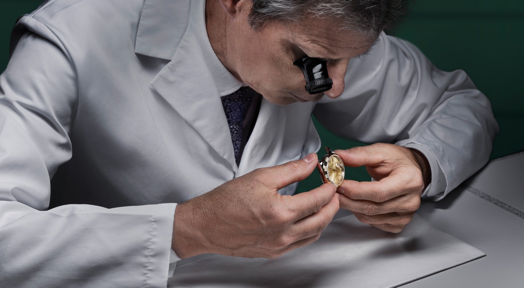 Rolex-trained expert male watchmaker in white collared coat and tie examines Rolex watch case without straps