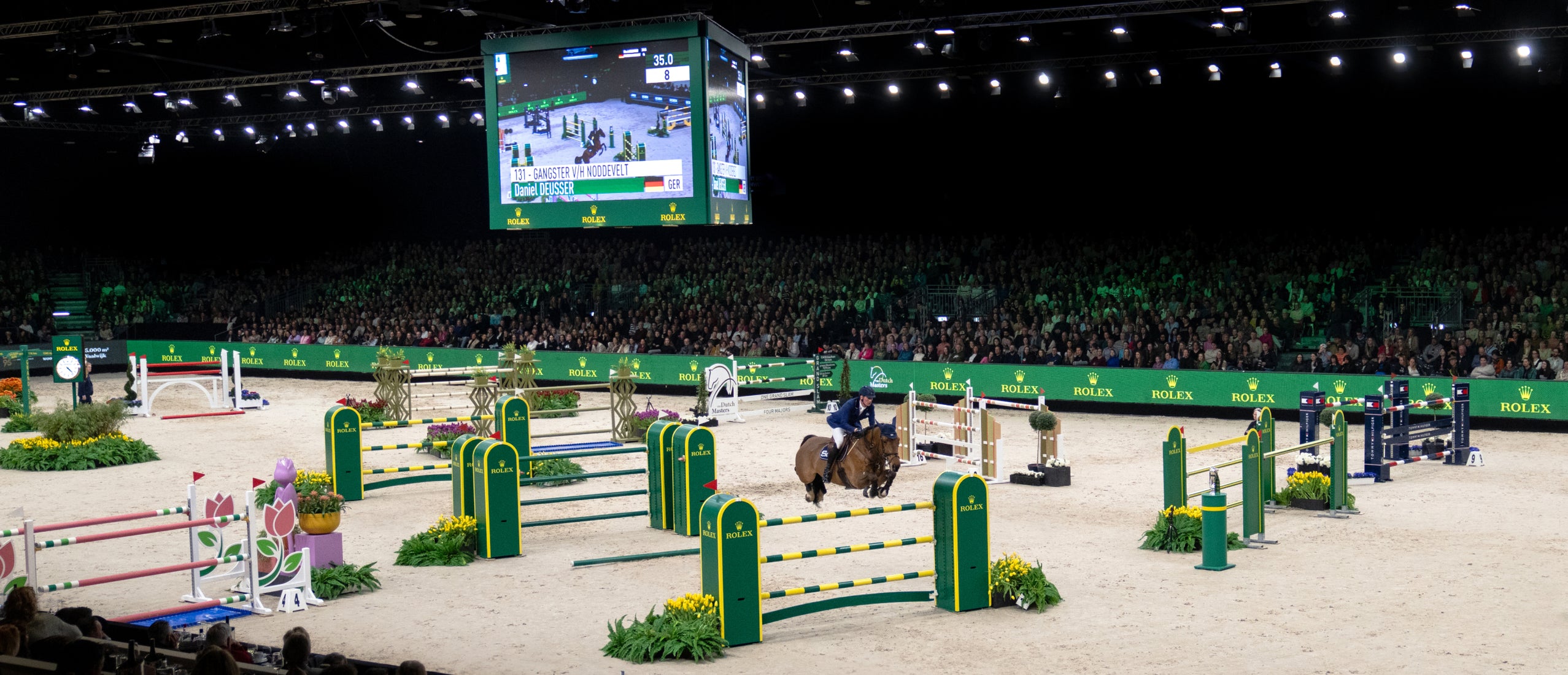 Indoor show jumping competition with a rider on a horse clearing a jump, surrounded by multiple colorful obstacles and Rolex branding, with a large audience and overhead display screen in the background.