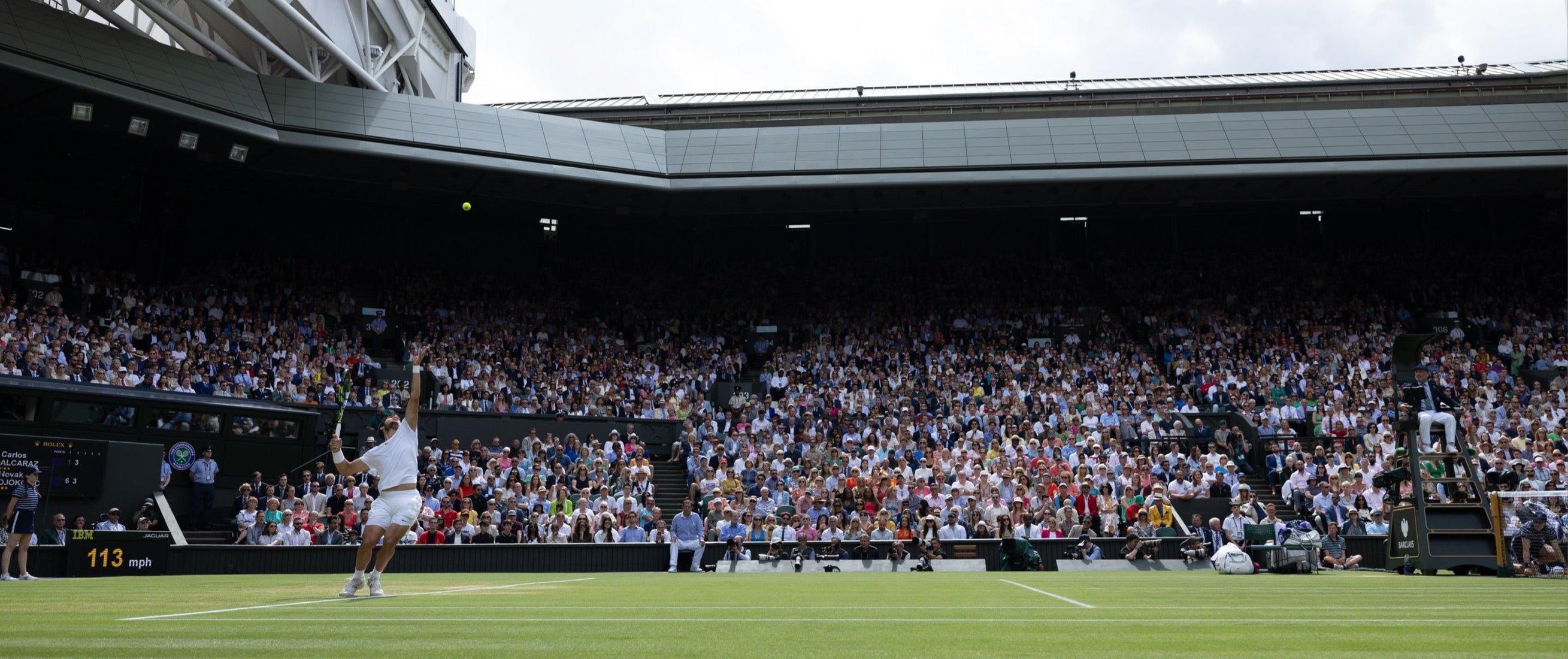 Wide-angle view of a grass tennis court at Wimbledon, with a player serving in front of a packed stadium filled with spectators.