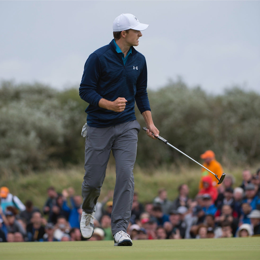 Golfer celebrating on the green with a fist pump, holding a putter, as crowds watch in the background during an outdoor golf tournament.