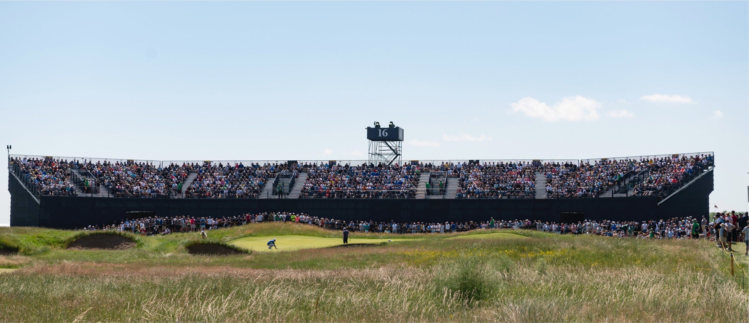 Large grandstand filled with spectators behind the 16th hole at a major golf championship, surrounded by tall grass and blue sky.