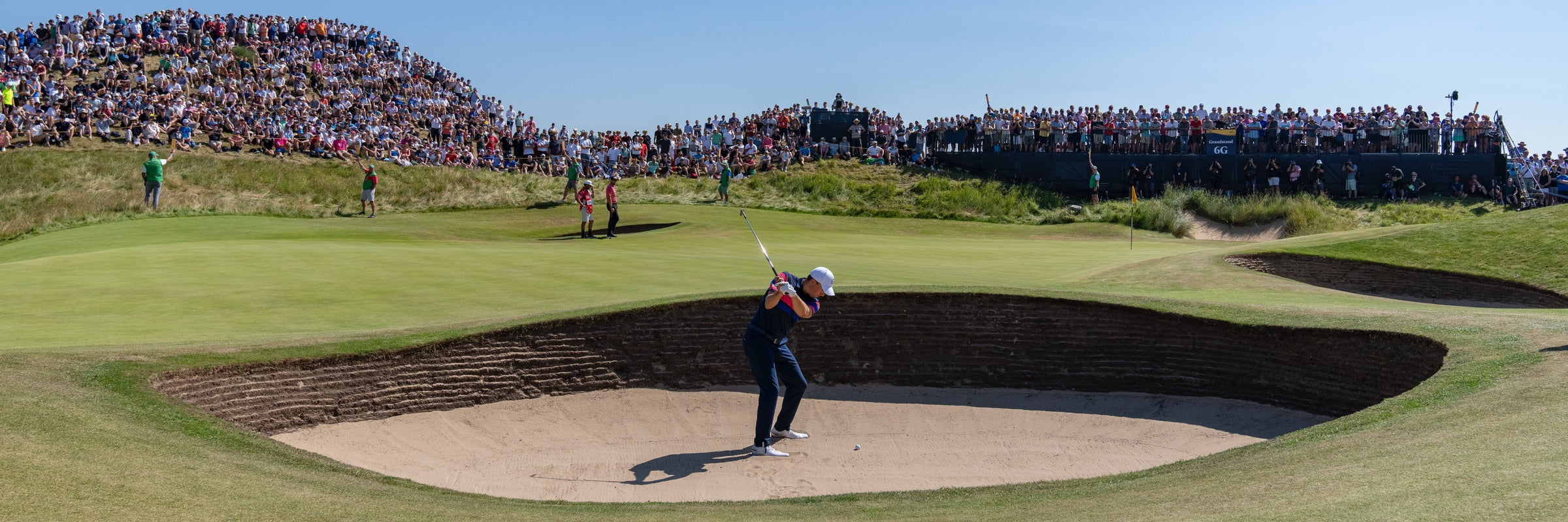 Golfer hitting a shot from a deep sand bunker towards the green, with large crowds watching from grassy mounds at a links-style golf course.