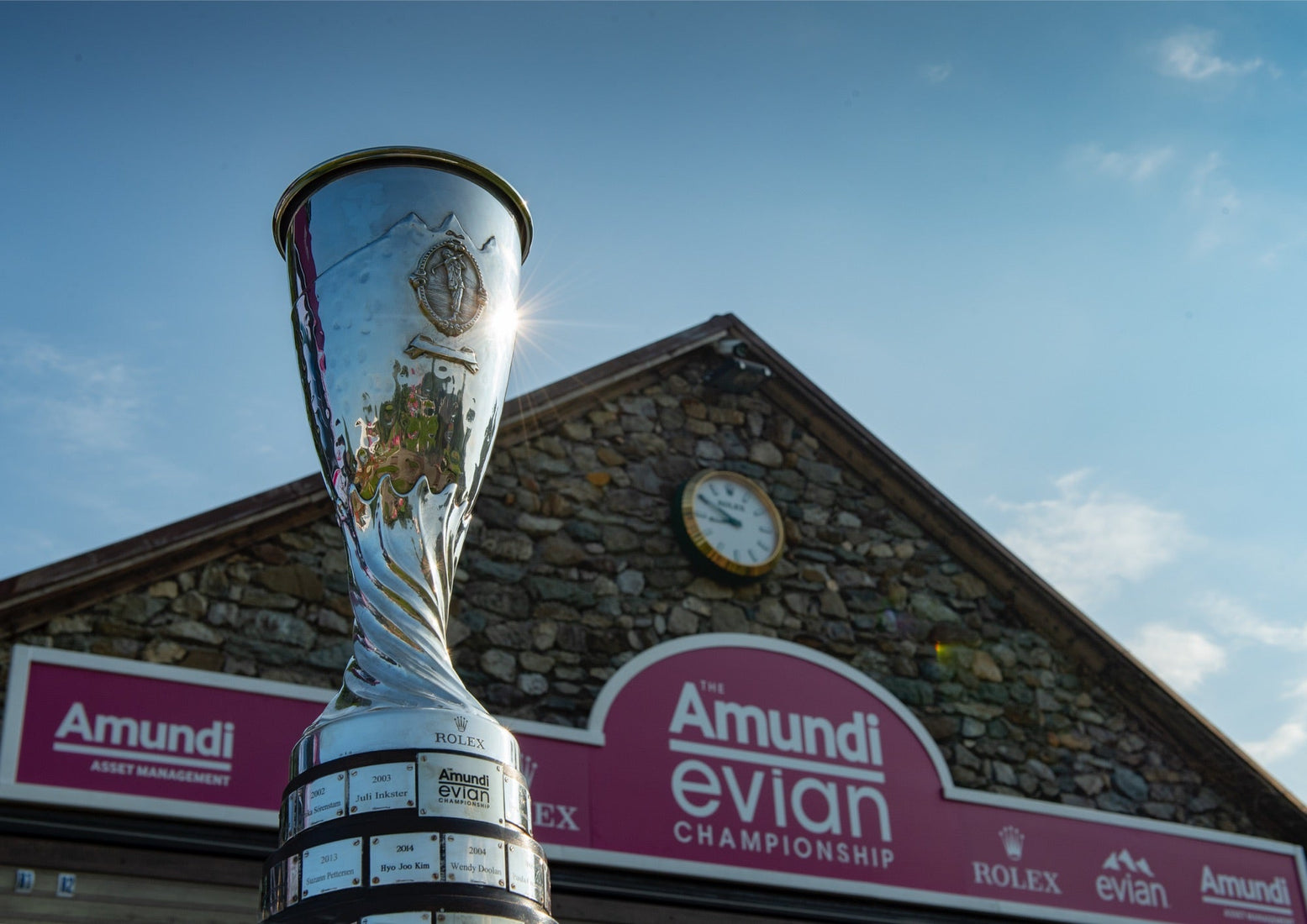 Silver Amundi Evian Championship trophy shining in the sunlight in front of a stone clubhouse with a Rolex clock and event signage under a clear blue sky.