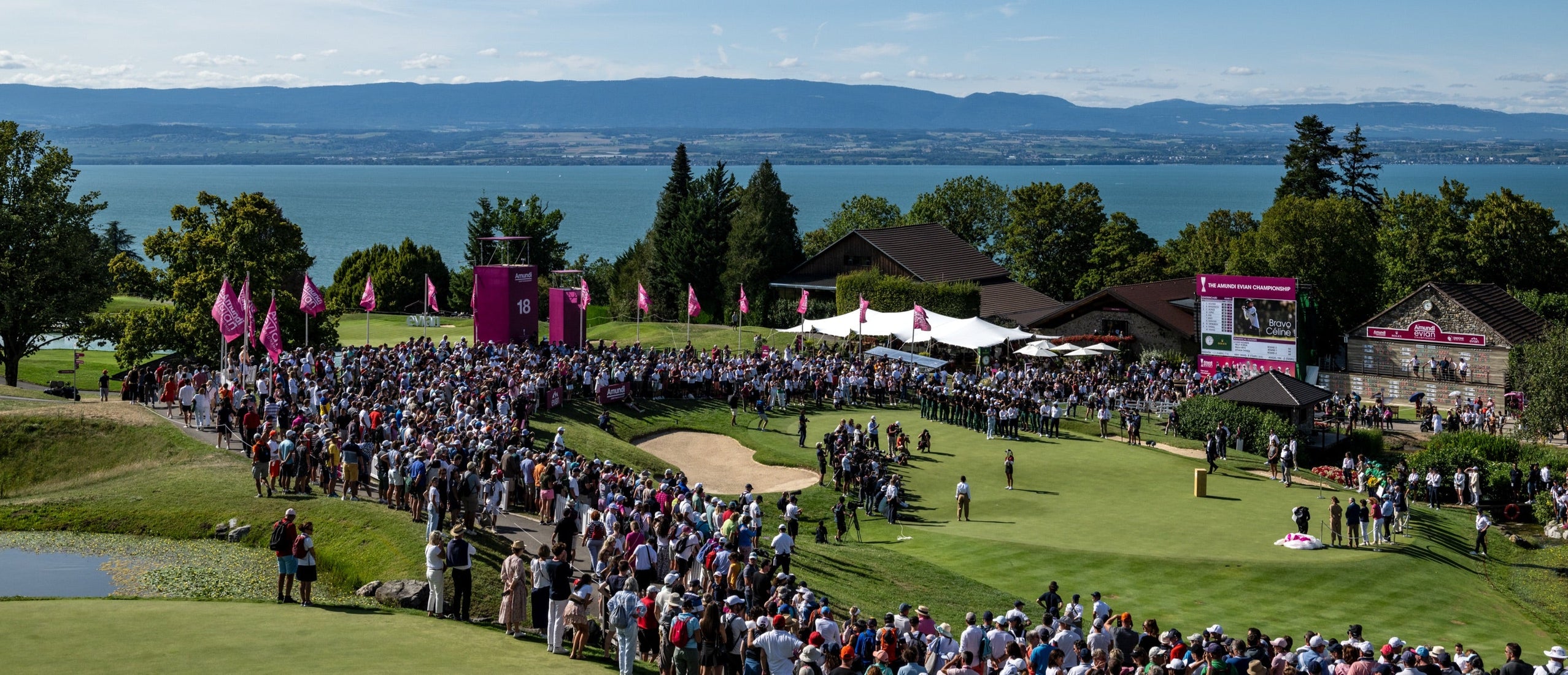 Large crowd gathered around the 18th green at a golf course overlooking a scenic lake and mountains during the Amundi Evian Championship, with event banners and scoreboards visible.