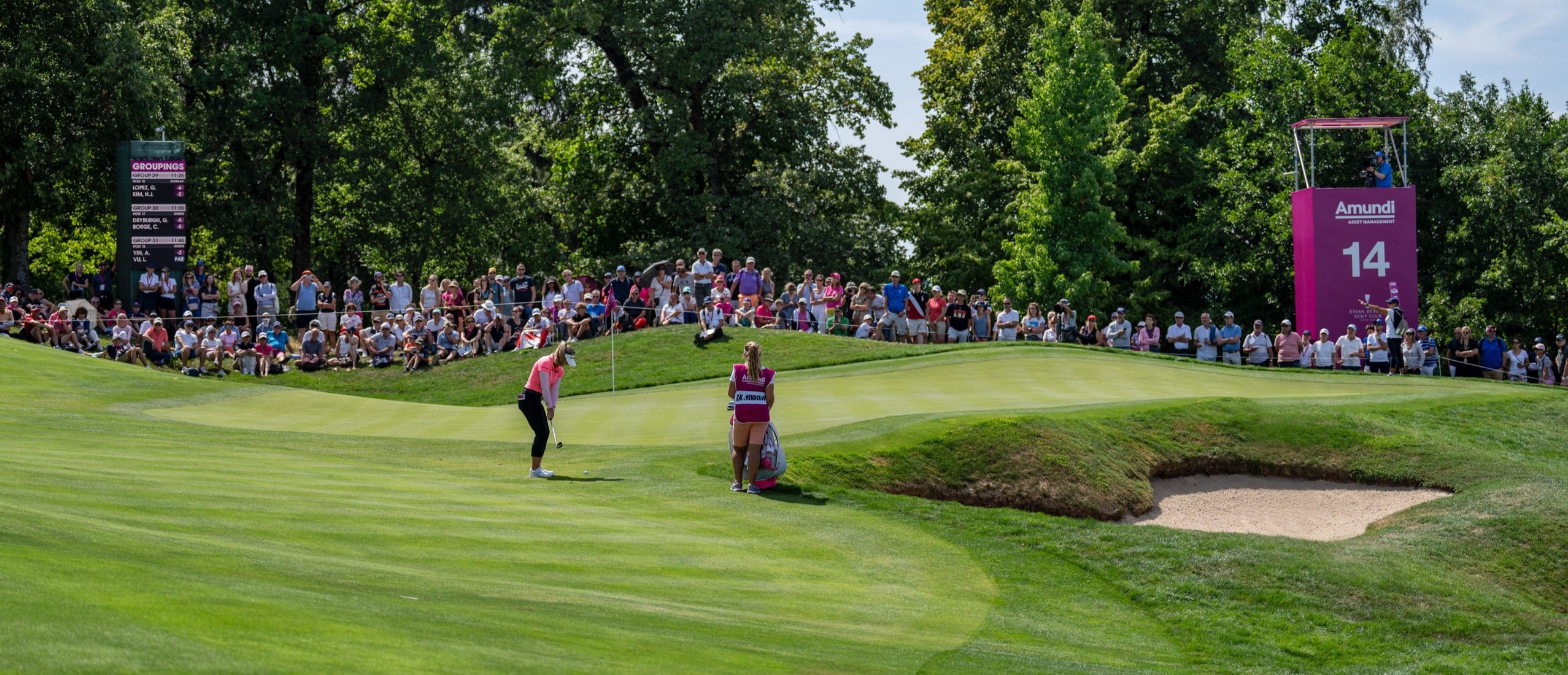 Golfer preparing to chip onto the green near a sand bunker on the 14th hole, surrounded by a gallery of spectators at an outdoor golf tournament.