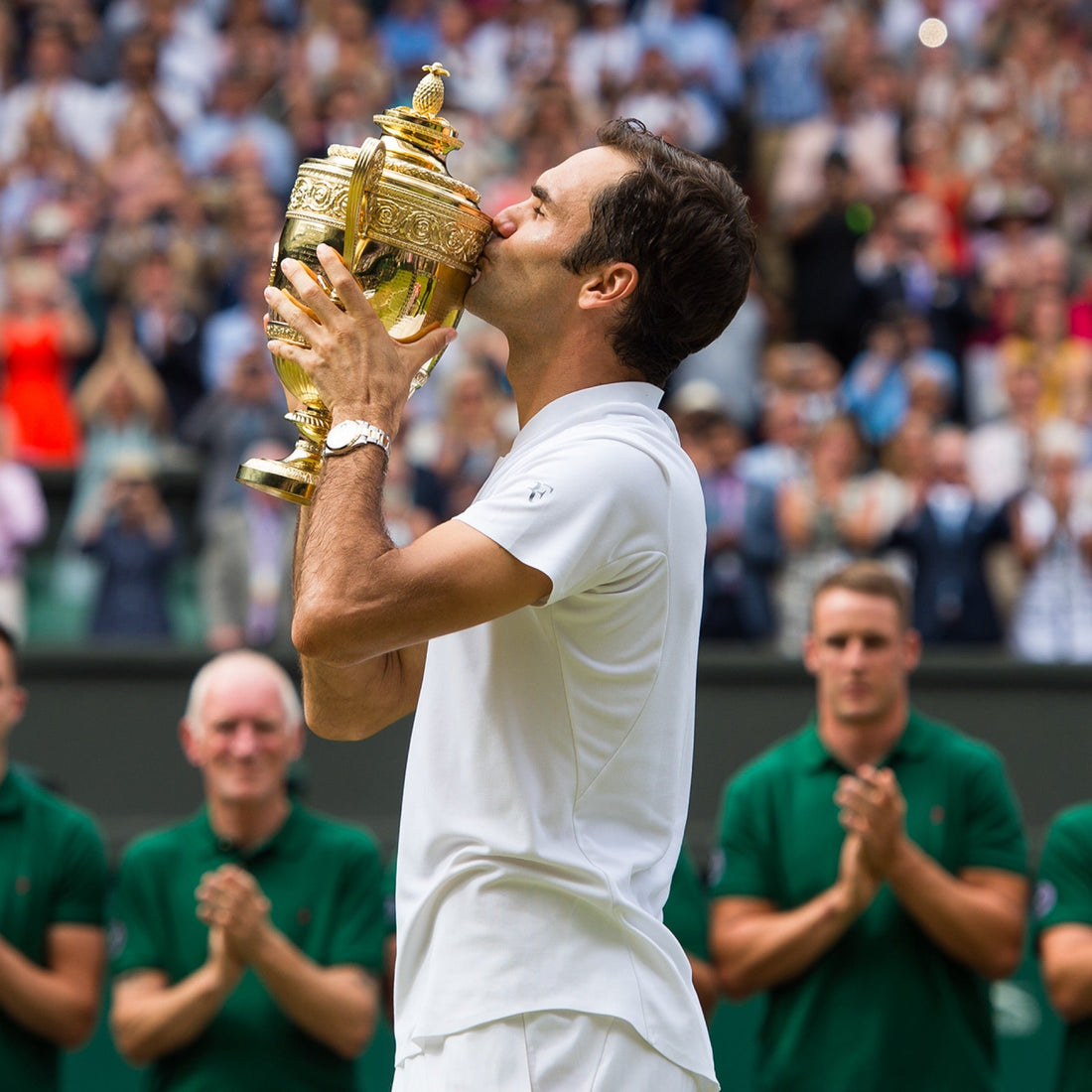 Tennis player in white kissing the Wimbledon gentlemen’s singles trophy on court, surrounded by applauding officials and a cheering crowd.